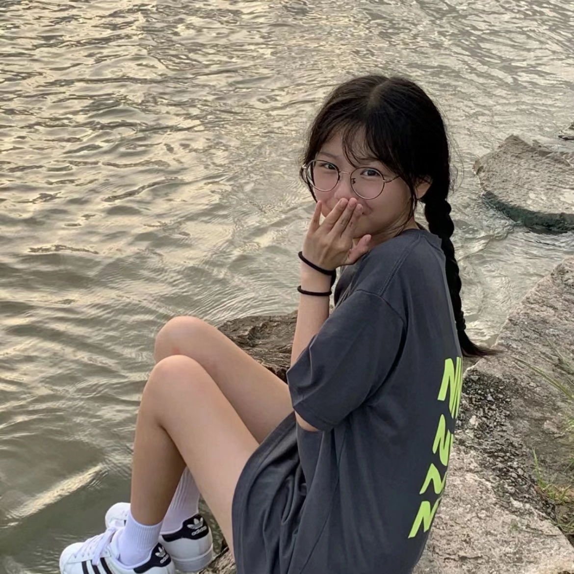 一位戴着眼镜和灰色衬衫的女人坐在水边的岩石上。