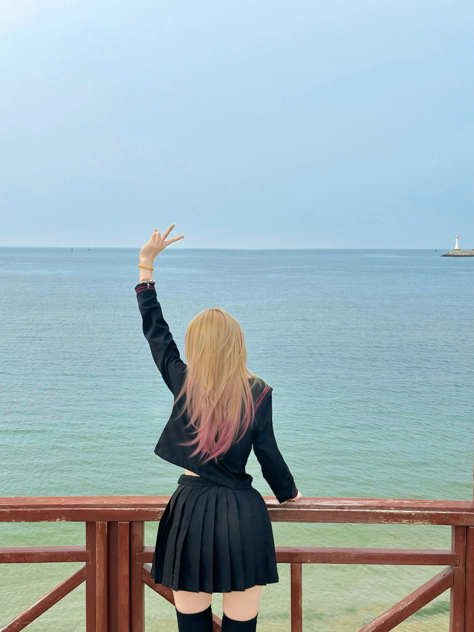 粉红色的头发和黑色夹克的女人在海洋上做和平的信号。