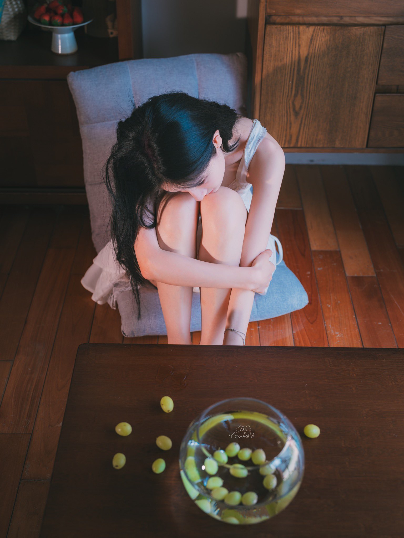 女人坐在沙发上,手臂围着腿,一碗葡萄在她面前。