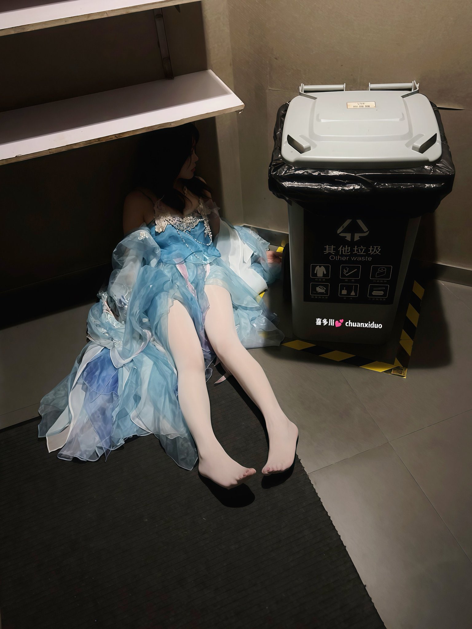 一位穿着蓝色礼服的女人躺在地板上,旁边是垃圾桶。