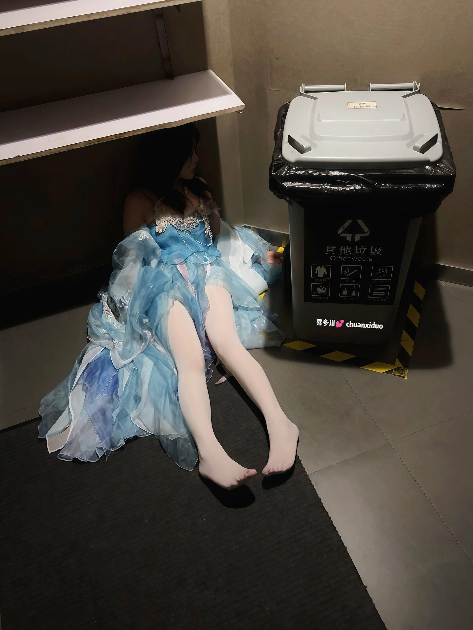 一位穿着蓝色礼服的女人躺在地板上,旁边是垃圾桶。