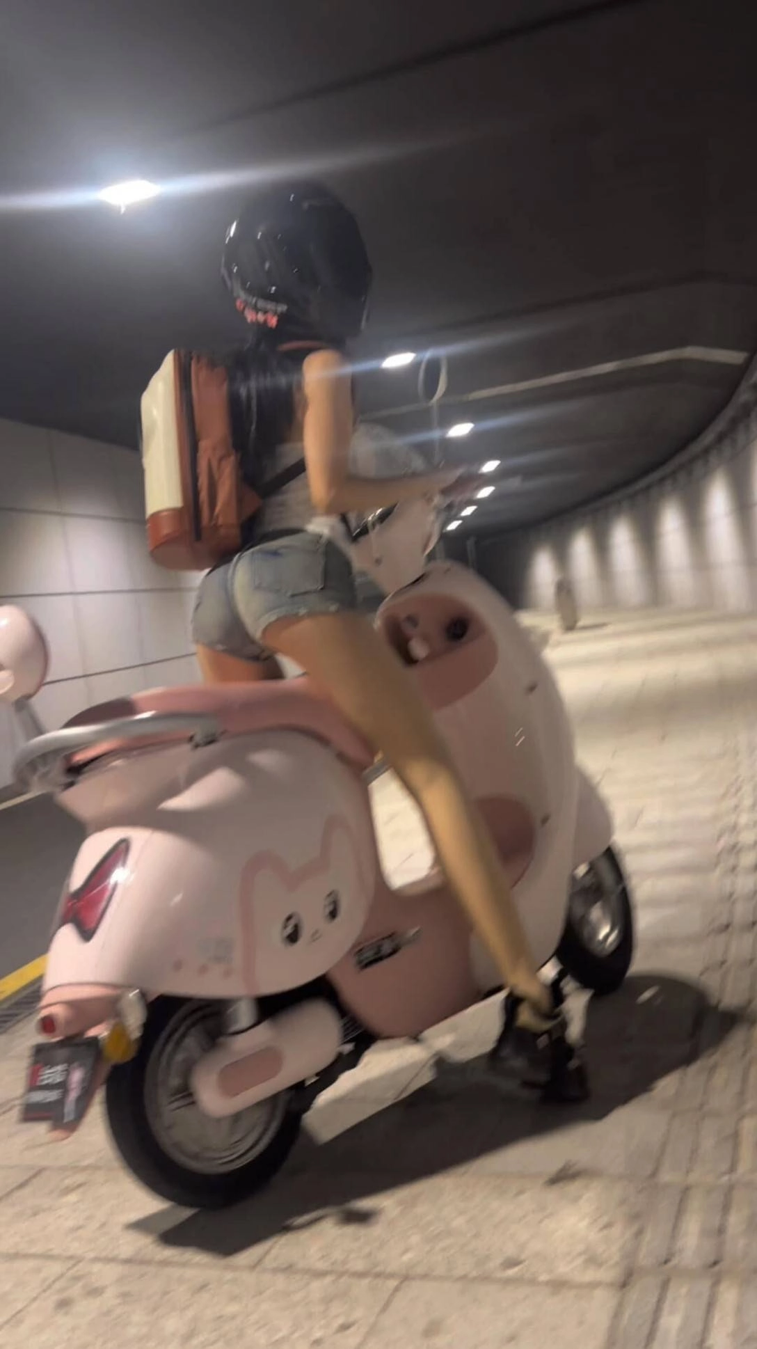 女人骑着一个粉红色的摩托车,背上有个袋子。